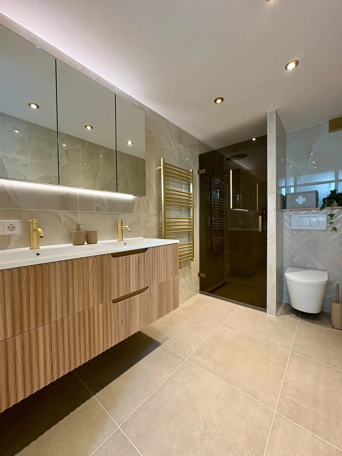 Luxe badkamer met eiken dubbele wastafel, gouden kranen, glazen douche en een visgraatwand bij het toilet